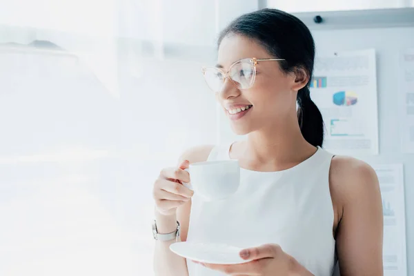 Hermosa mujer de negocios latina sosteniendo la taza de café y mirando hacia otro lado en la oficina - foto de stock