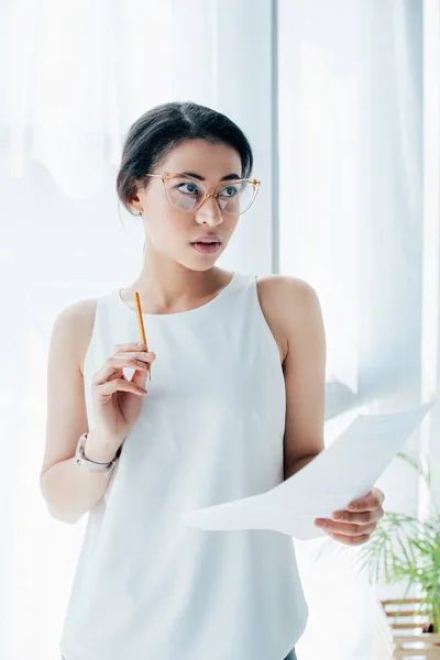 Nachdenkliche Geschäftsfrau hält Papier und Bleistift in der Hand, während sie im Büro wegschaut — Stockfoto