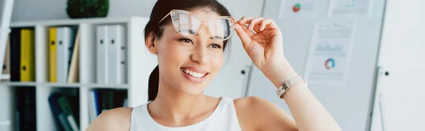 Plan panoramique de femme d'affaires latine gaie touchant des lunettes et regardant loin dans le bureau — Photo de stock