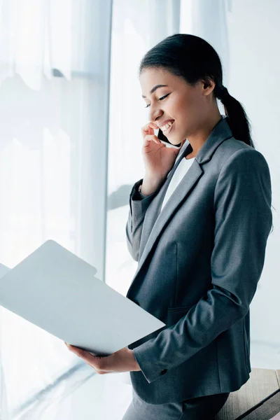 Cheerful latin businesswoman talking on smartphone while looking at documents in paper folder — Stock Photo