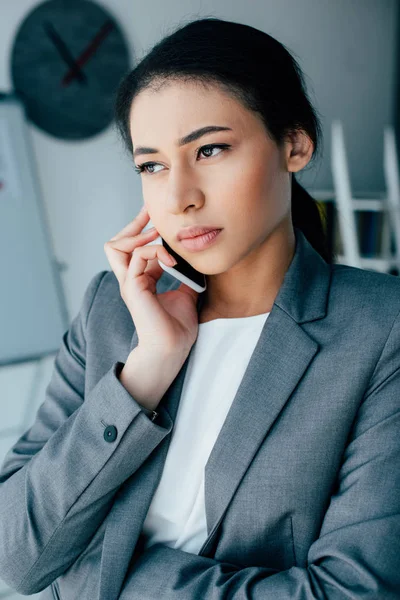 Mujer de negocios latina hablando en un teléfono inteligente mientras mira hacia otro lado en la oficina - foto de stock