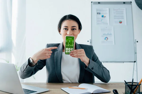 Schöne lateinische Geschäftsfrau zeigt mit dem Finger auf Smartphone mit Herzschlagrate auf dem Bildschirm — Stockfoto