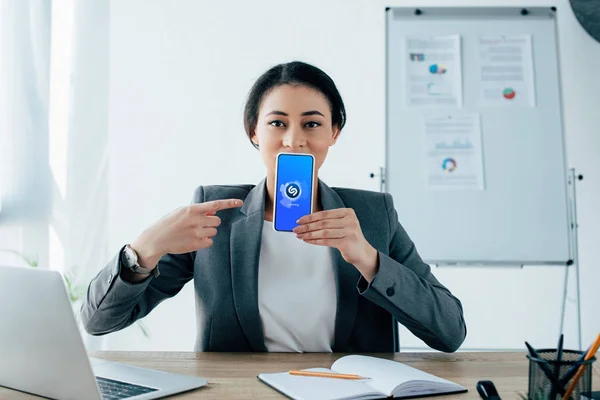 KYIV, UCRANIA - 26 DE ABRIL DE 2019: Hermosa mujer de negocios latina mostrando el teléfono inteligente con la aplicación Shazam en la pantalla . - foto de stock