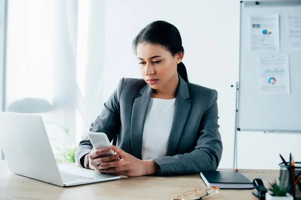 Serious latin businesswoman using smartphone while sitting at workplace near laptop — Stock Photo