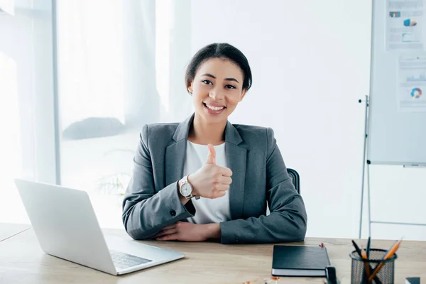 Belle femme d'affaires latine montrant pouce vers le haut tout en étant assis sur le lieu de travail près d'un ordinateur portable et souriant à la caméra — Photo de stock