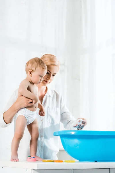 Low angle view of attractive woman holding toddler son while standing near blue baby bathtub — Stock Photo