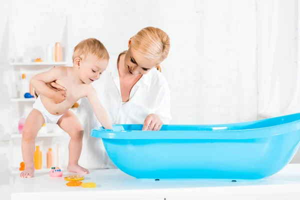 Happy blonde mother looking at baby bathtub and holding cute toddler son in bathroom — Stock Photo