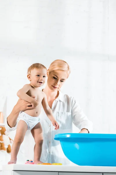 Vista angolo basso di madre attraente che tiene felice figlio bambino vicino vasca da bagno bambino — Foto stock