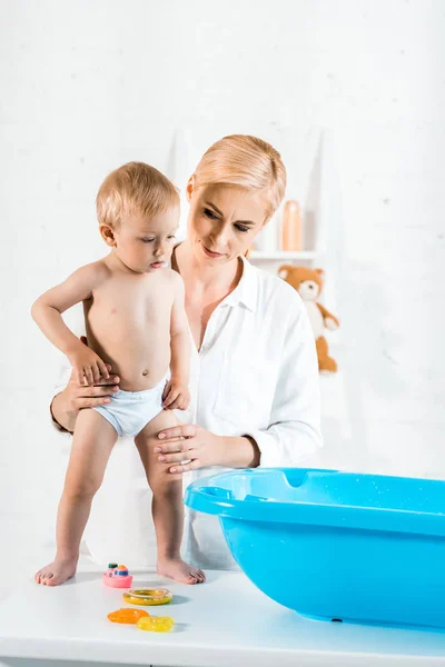 Schöne Mutter schaut auf Babybadewanne, während sie ihren kleinen Sohn im Badezimmer hält — Stockfoto