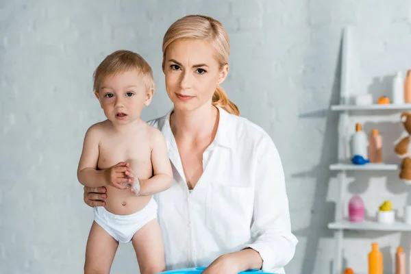 Blonde femme regardant caméra près mignon tout-petit fils à la maison — Photo de stock