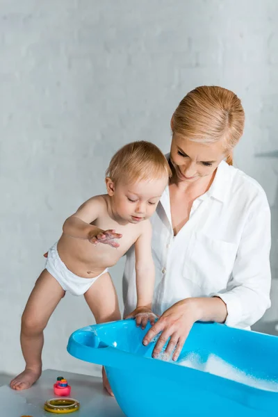 Mãe loira de pé perto do filho da criança olhando para banheira de bebê azul — Fotografia de Stock