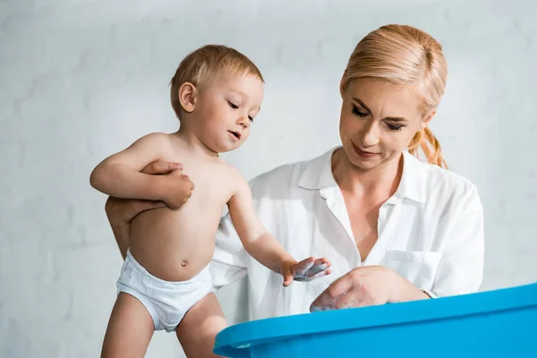 Low angle view of blonde woman standing and looking at blue baby bathtub near toddler son — Stock Photo
