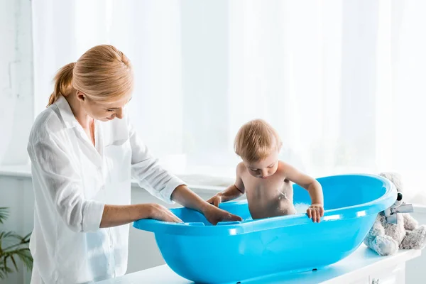 Glückliche Mutter, die neben niedlichem Kleinkind-Sohn steht und zu Hause baden geht — Stockfoto