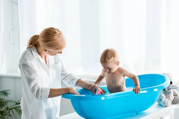 Blonde mother standing near cute toddler son taking bath at home — Stock Photo