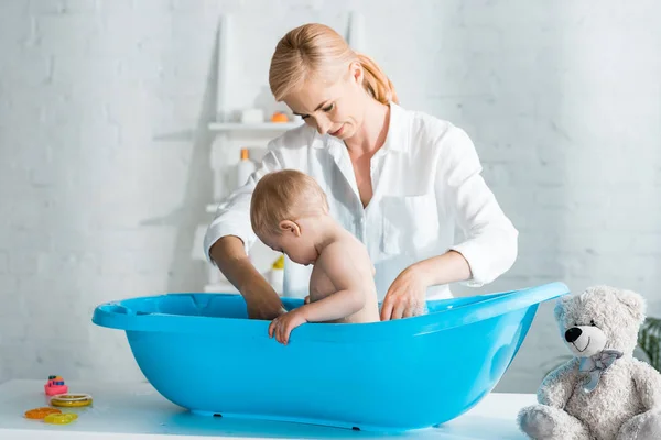 Beautiful mother standing near toddler kid in baby bathtub — Stock Photo