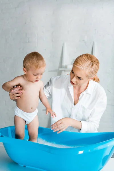 Beautiful mother looking at cute toddler kid in baby bathtub — Stock Photo