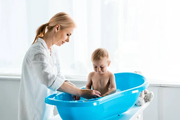 Bonito criança tomando banho perto feliz loira mãe em casa — Fotografia de Stock