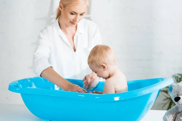 Cheerful blonde mother smiling near cute toddler son taking bath — Stock Photo