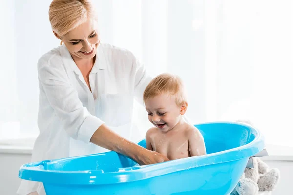 Cheerful blonde mother smiling while washing cute toddler son in blue baby bathtub — Stock Photo