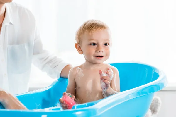 Cropped view of mother washing happy toddler son in blue baby bathtub — Stock Photo