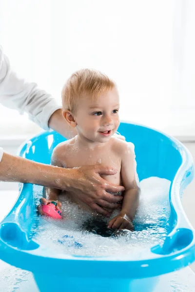 Abgeschnittene Ansicht der Mutter, die fröhliches Kleinkind in blauer Babybadewanne wäscht — Stockfoto