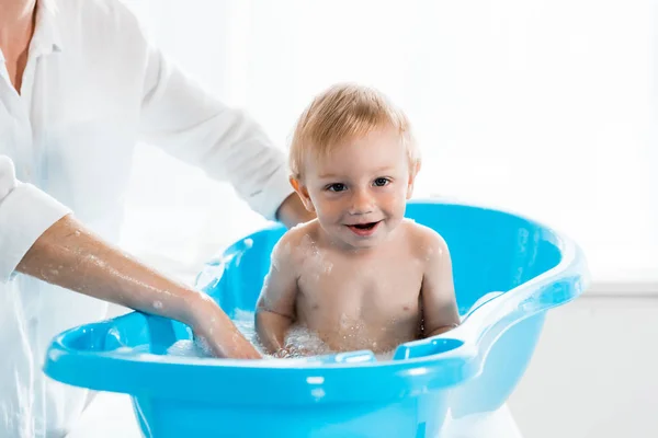 Обрізаний вид мами, що миє щасливу дитину у блакитній дитячій ванні — стокове фото