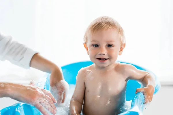 Vista recortada de la madre cerca del niño feliz en la bañera azul del bebé - foto de stock