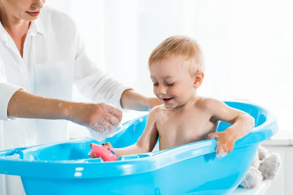 Vista recortada de la madre feliz cerca del niño alegre en la bañera azul del bebé - foto de stock