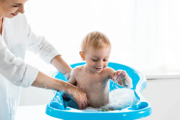 Vue recadrée de mère heureuse lavant tout-petit enfant dans la baignoire bleue pour bébé — Photo de stock