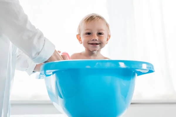 Vue recadrée de la mère laver heureux tout-petit enfant dans la baignoire bleue bébé — Photo de stock