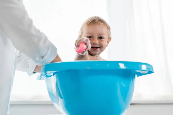 Vista recortada de la madre lavando niño feliz señalando con el dedo en la bañera azul del bebé - foto de stock