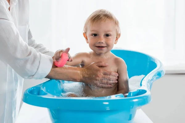 Vue recadrée de la mère lavant heureux tout-petit enfant avec jouet en caoutchouc dans la baignoire bleue pour bébé — Photo de stock