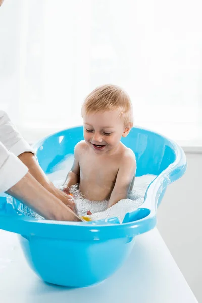Cropped view of mother washing smiling toddler child in blue baby bathtub — Stock Photo