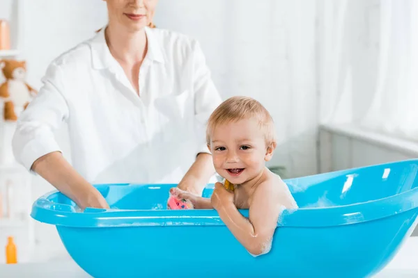 Selective focus of cute smiling toddler boy in baby bathtub near mother — Stock Photo