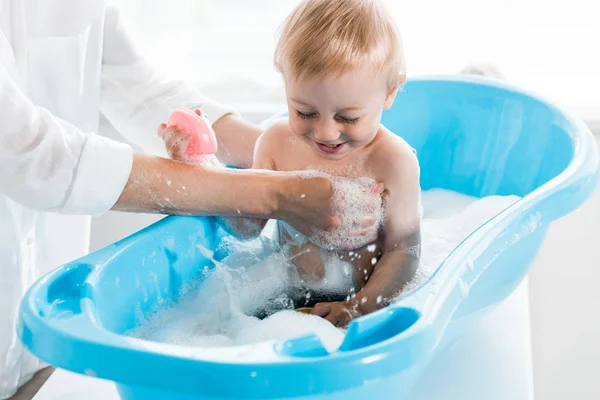 Cropped view of mother washing smiling toddler kid in blue baby bathtub — Stock Photo