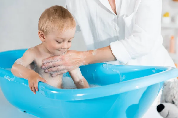Обрезанный вид на мать стирает милый малыш сын в голубой ванне ребенка — стоковое фото
