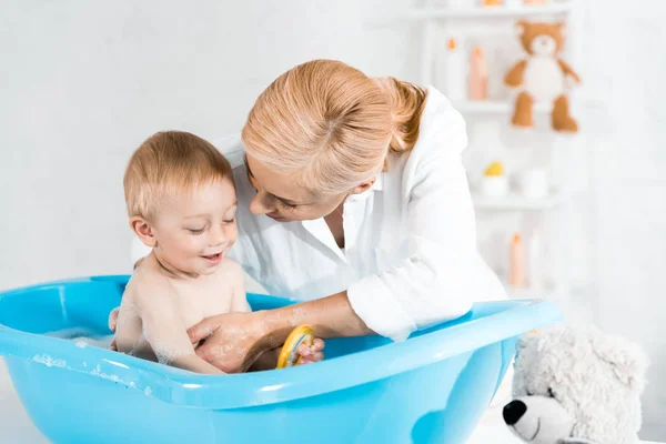 Blonde Mutter schaut Kleinkind in blauer Babywanne lächelnd an — Stockfoto