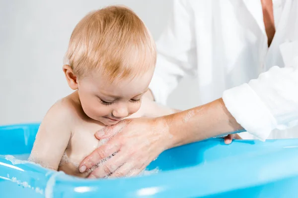 Recortado vista de la mujer lavado lindo niño pequeño en bañera de bebé - foto de stock