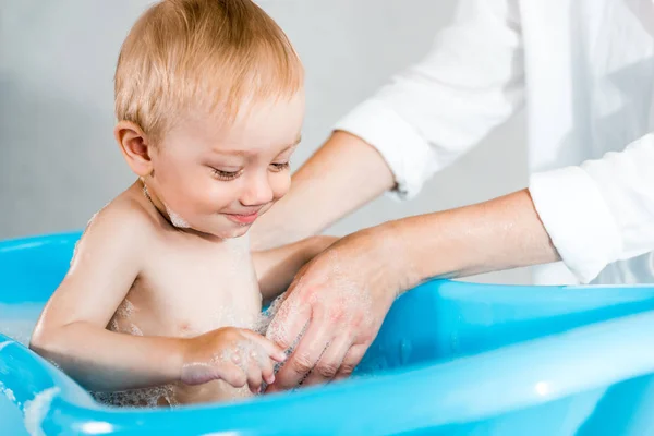 Recortado vista de la mujer lavado lindo niño pequeño en bañera de bebé - foto de stock