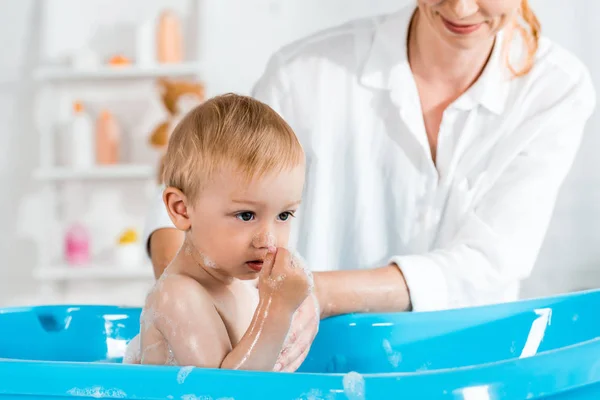 Vista recortada de la mujer sonriente lavando niño pequeño en bañera de bebé - foto de stock