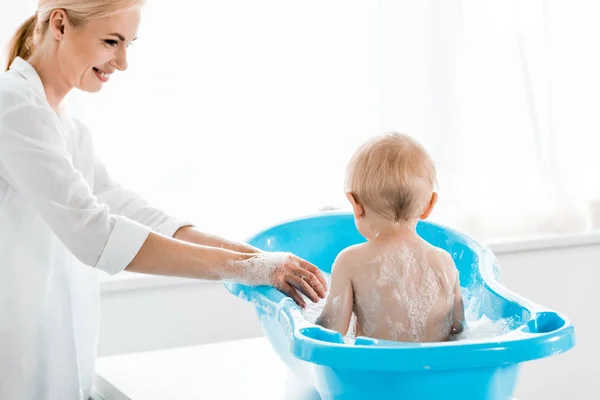 Привлекательная и улыбающаяся мать стирает малыша сына в голубой ванне ребенка — стоковое фото