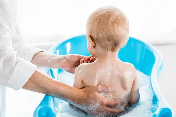 Vista recortada de la mujer lavando niño pequeño en bañera de bebé - foto de stock