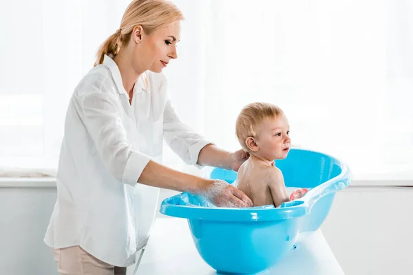 Attractive mother washing toddler son in blue plastic baby bathtub — Stock Photo