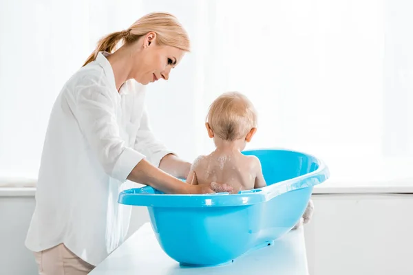 Atractiva madre lavando niño pequeño en azul bañera de plástico bebé - foto de stock