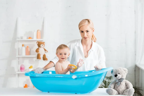 Счастливая мать улыбается рядом с малышом сыном в пластиковой ванне ребенка — стоковое фото