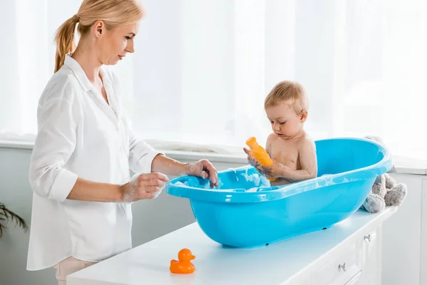 Madre rubia mirando a su hijo pequeño sosteniendo botella con champú - foto de stock