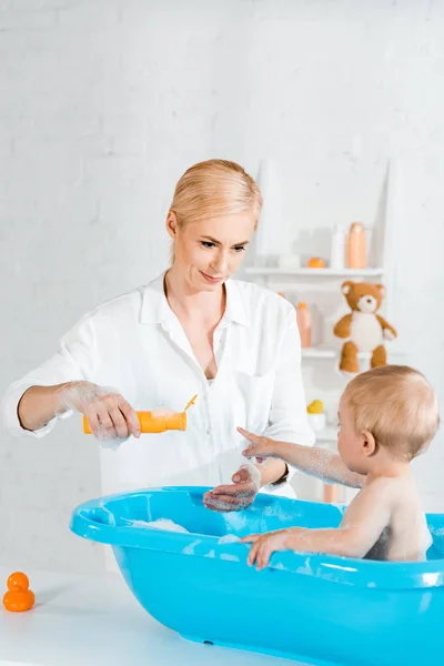 Lindo niño apuntando con el dedo a la botella con champú cerca de la madre - foto de stock