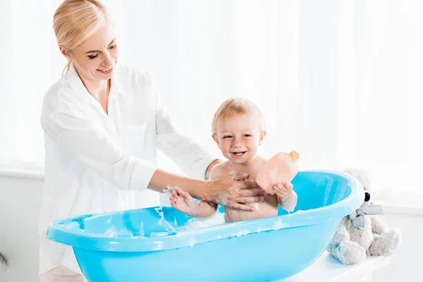 Mãe feliz lavar criança alegre filho segurando garrafa com xampu — Fotografia de Stock