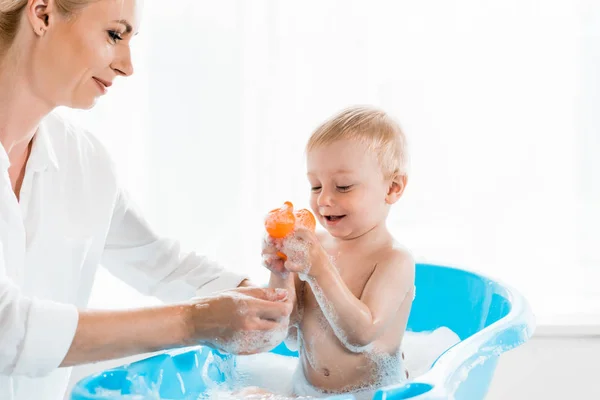 Mãe feliz lavar criança alegre filho segurando pato de borracha no banheiro — Fotografia de Stock