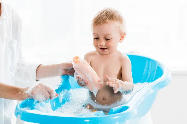 Обрізаний вид матері біля веселого маленького сина, що тримає пляшку з шампунем у ванній — стокове фото